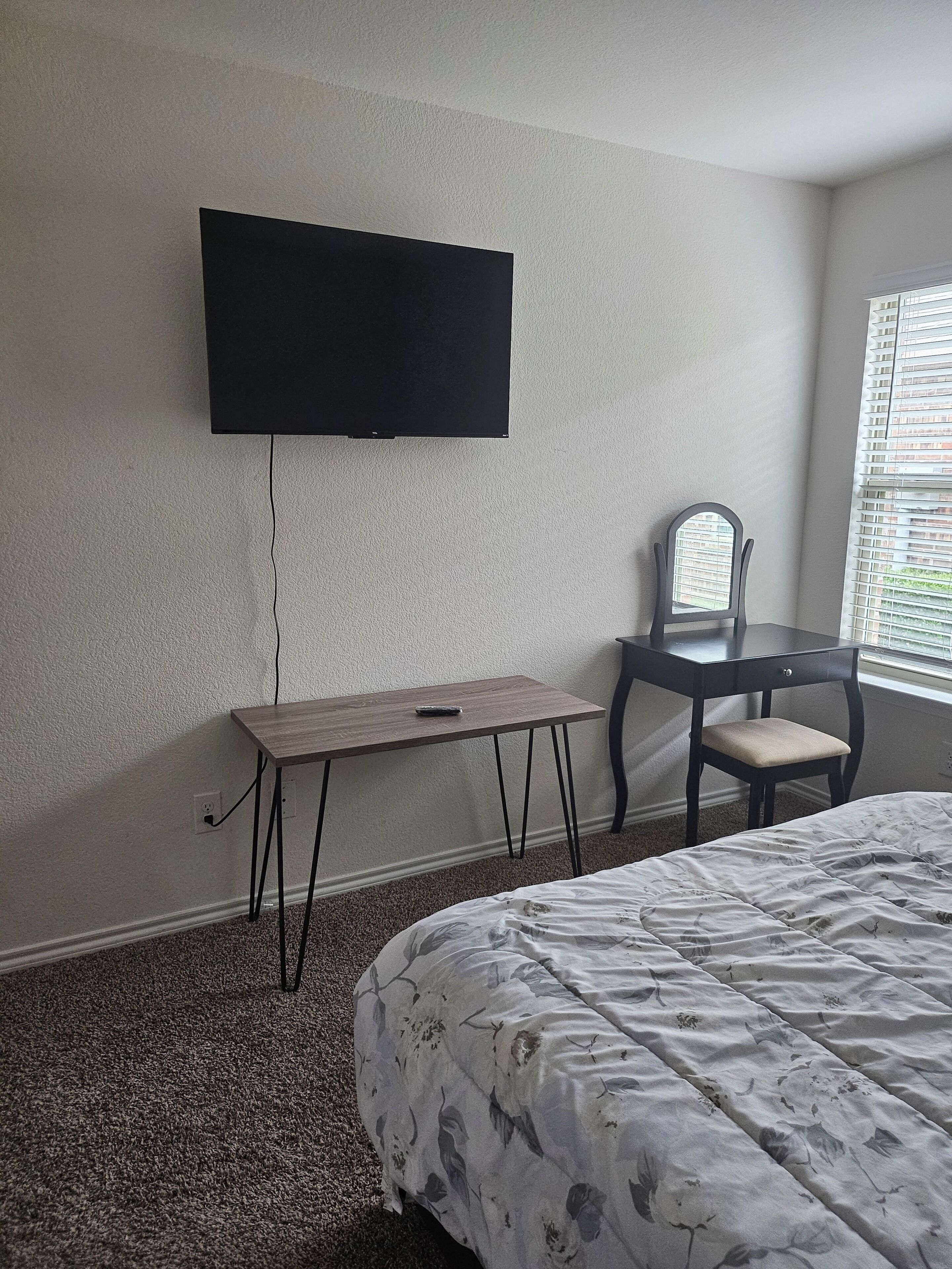 Ein Zimmer mit einem Bett, einem Schreibtisch, einem Sessel und einem an der Wand befestigten Fernseher.