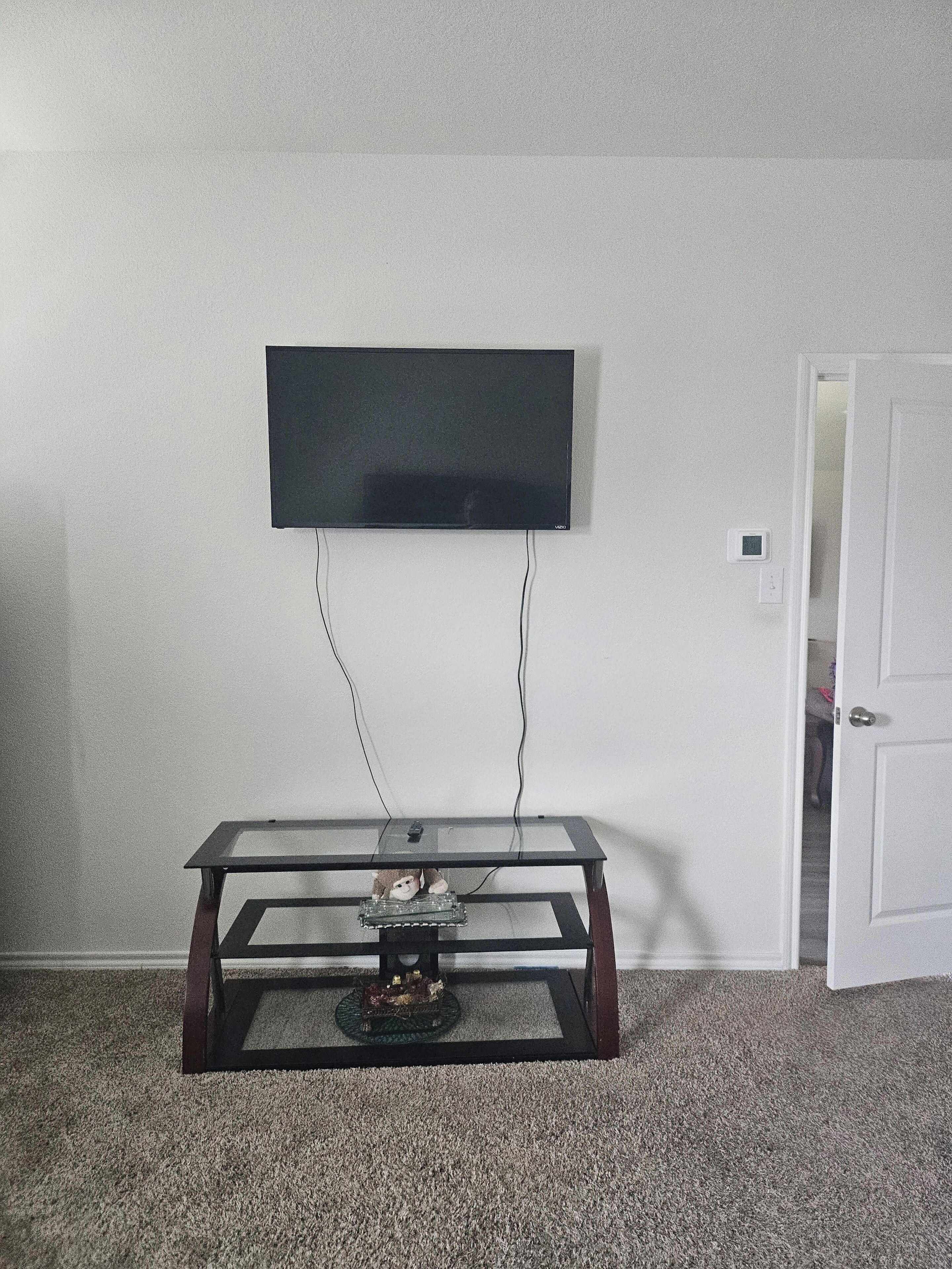 Ein Zimmer mit an der Wand montiertem Fernseher, einem Glastisch mit Fernbedienung und einer geschlossenen Tür.