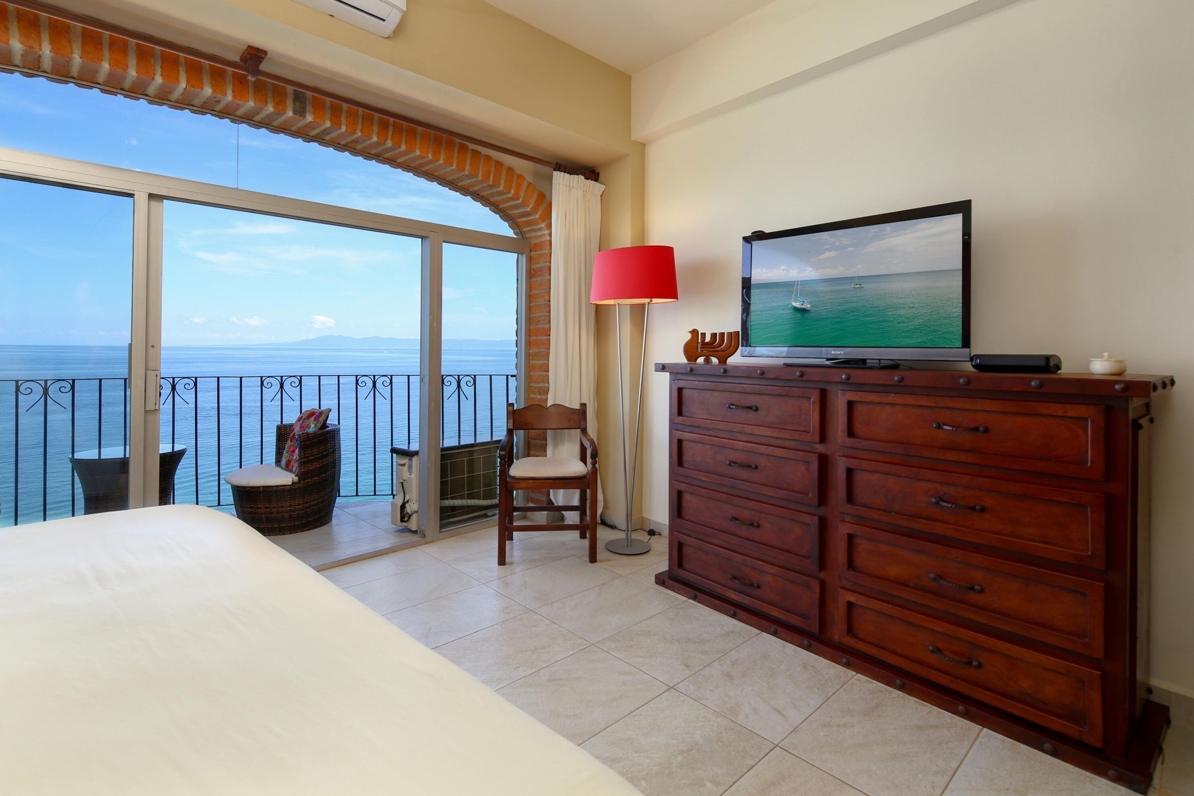 Una habitación de hotel con balcón, una cama, un aparador de madera, un televisor y una silla.