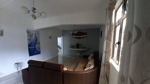 Un salon avec un canapé en osier marron, un mur blanc orné d’un tableau représentant un paon, un ventilateur de plafond et une fenêtre avec des rideaux à motifs.
