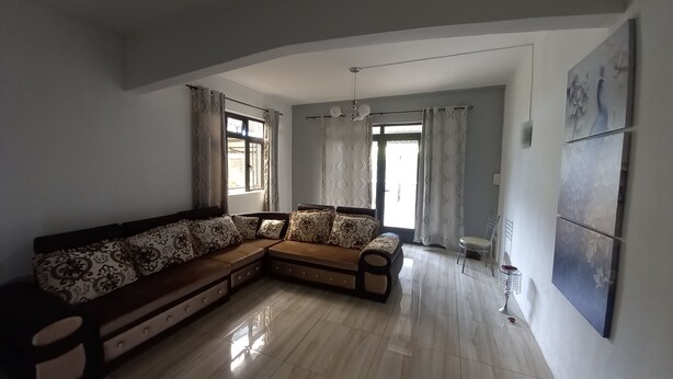 Un salon moderne comprenant un canapé d’angle, une table à manger et une chaise. Il y a deux fenêtres avec des rideaux, un ventilateur de plafond et un luminaire fixé au mur.