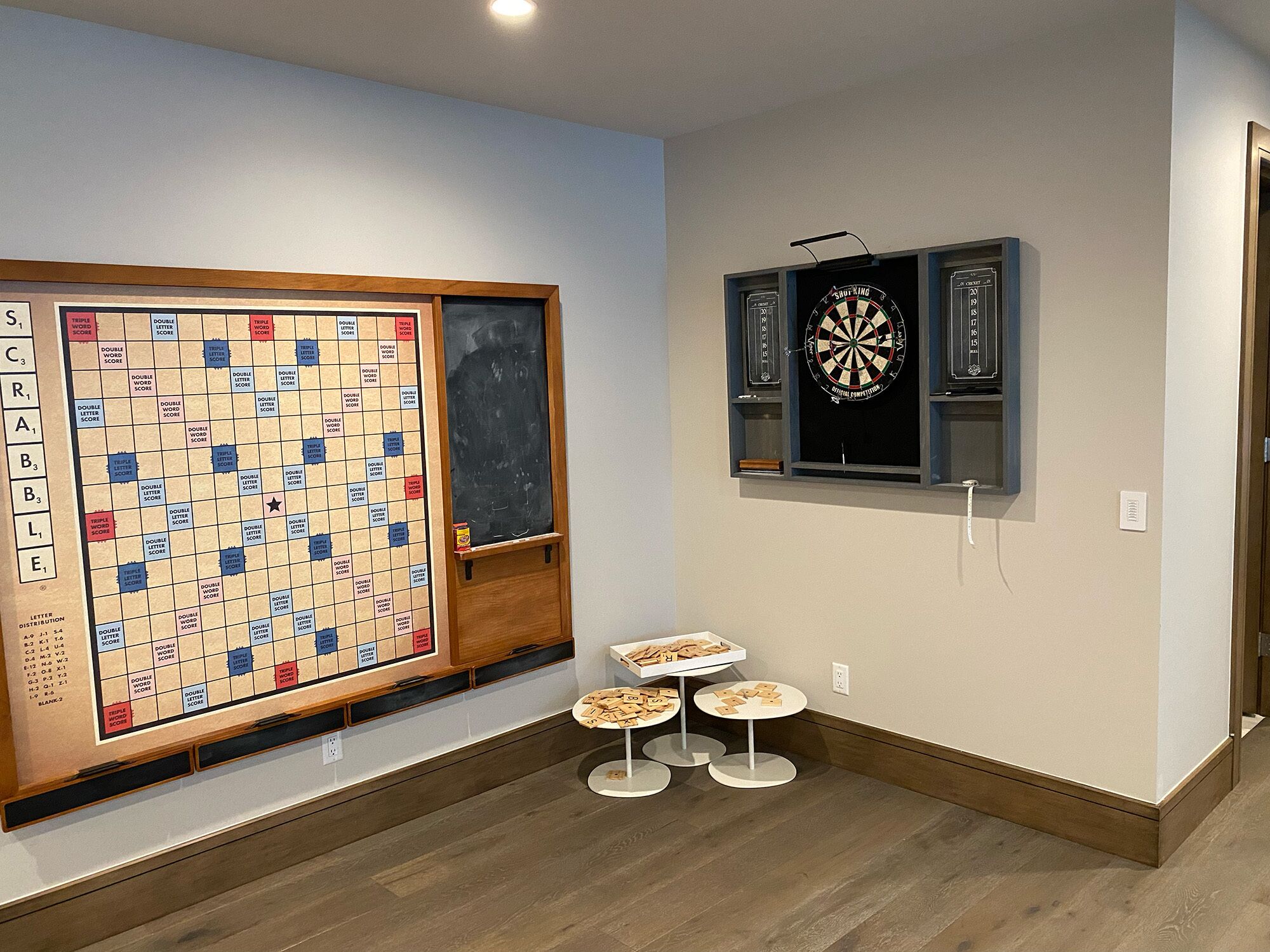 Una habitación con un tablero de Scrabble, un dardo y dos mesitas con fichas.