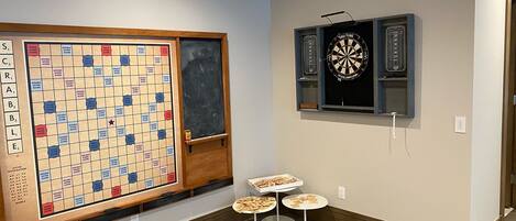 Una habitación con un tablero de Scrabble, un dardo y dos mesitas con fichas.