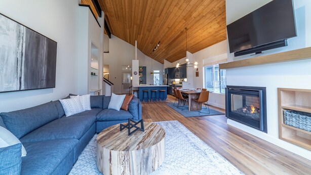 A modern living room with a blue sectional sofa, a round coffee table, a fireplace, and a kitchen area in the background. - Snowcreek Luxury Remodel 529 (Mammoth Lakes)
