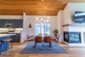 A modern kitchen with grey cabinets, a microwave, and a stove. There is a dining area with a wooden table and four chairs. A large window provides natural light, and a fireplace is visible on the right. - Snowcreek Luxury Remodel 529 (Mammoth Lakes)