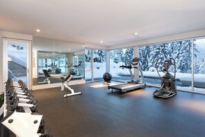 A modern gym with exercise equipment, large windows, and a view of snow-covered trees. - Kadenwood 2980 - Ski in Ski out, Private Pool, Hot Tub & Gondola (Whistler)