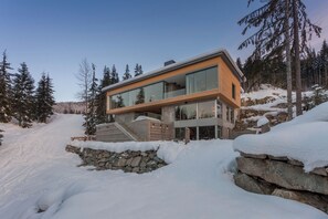A modern house with large glass windows, a stone foundation, and a wooden deck.