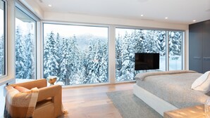 A modern bedroom with large windows, a bed, a TV, and a cozy seating area. - Kadenwood 2980 – Ski in Ski out, Private Pool, Hot Tub & Gondola (Whistler)