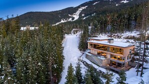 A modern, multi-story house surrounded by snow-covered trees and a mountain backdrop. - Kadenwood 2980 – Ski in Ski out, Private Pool, Hot Tub & Gondola (Whistler)