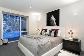 A modern bedroom with a large bed, a nightstand, a wall-mounted lamp, and a window with a view of a snowy landscape. - Kadenwood 2980 - Ski in Ski out, Private Pool, Hot Tub & Gondola (Whistler)