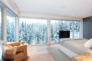 A modern bedroom with large windows, a bed, a TV, and a cozy seating area.