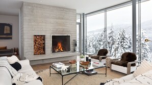A modern living room with a fireplace, large windows, and a view of snow-covered trees. - Kadenwood 2980 – Ski in Ski out, Private Pool, Hot Tub & Gondola (Whistler)