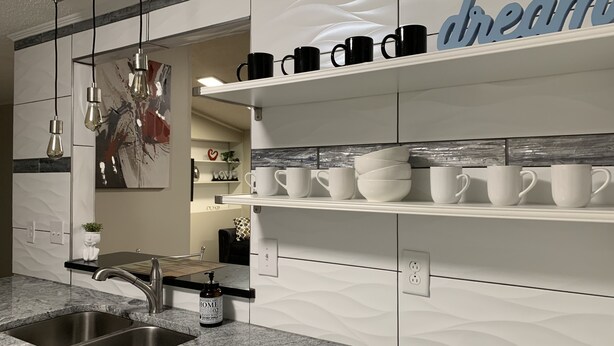 Cocina moderna con encimera de granito, pileta de acero inoxidable y muebles de cocina blancos. Hay estantes con tazas blancas y negras, y un cartel decorativo que dice 'dream'.