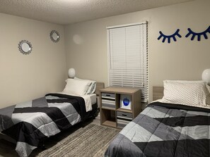 A bedroom with two beds, a nightstand, a window with blinds, and wall decorations.