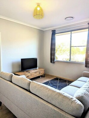 Ein modernes Wohnzimmer mit einer Couch, einem Fernseher auf einem Holzboden und einem Fenster mit Vorhängen.