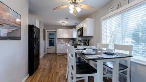 Une cuisine moderne avec des meubles blancs, un réfrigérateur noir, une table à manger avec des chaises blanches et un ventilateur de plafond.