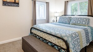 Une chambre à coucher avec un lit, une table de chevet, une lampe, une fenêtre, un ventilateur de plafond et un tableau au mur.