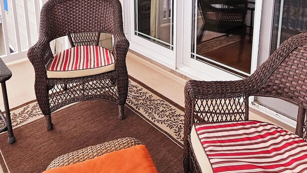 Une véranda couverte, aménagée avec des meubles en osier, notamment des chaises et un banc, et dotée d’une porte-fenêtre coulissante.