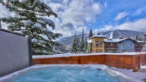 Un área de hidromasaje con terraza de madera, rodeada de árboles y edificios cubiertos de nieve.
