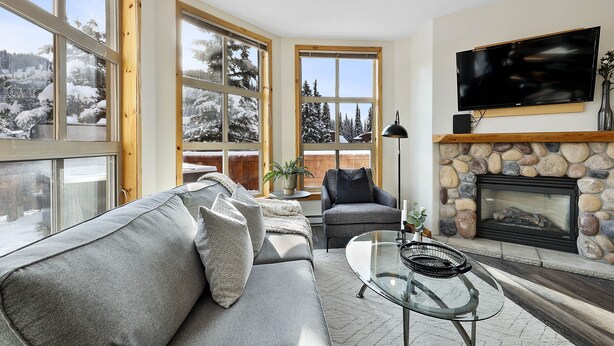Una sala de estar acogedora con chimenea de piedra, amplias ventanas con vistas nevadas, un sofá confortable y un televisor de pantalla plana.