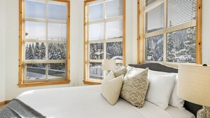 Un dormitorio con una cama grande, dos ventanas con persianas, una lámpara de noche y vistas a árboles cubiertos de nieve.