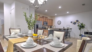 A modern dining area with a wooden table set for six, surrounded by white chairs. A large potted plant and a vase with flowers are on the table. - Casita Beth near Disney & Orlando Parks! (Kissimmee)