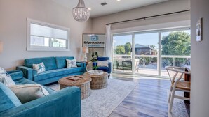A spacious living room with a blue sofa, wicker ottomans, and a chandelier. A sign for 'itripvacations key west' is visible on the wall. - The Salty Sailfish: Live the Keys Life, Stunning Canal Front Home, Pool, Dock & Dog-Friendly Bliss! (Summerland Key)