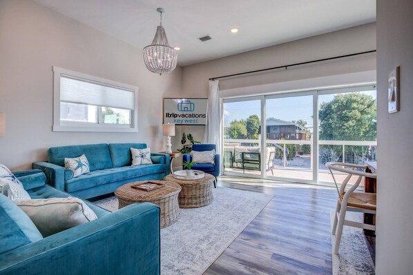 A spacious living room with a blue sofa, wicker ottomans, and a chandelier. A sign for 'itripvacations key west' is visible on the wall.