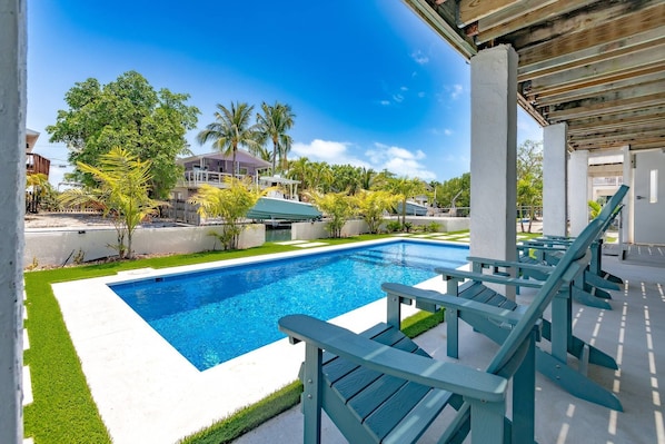 A swimming pool with a wooden deck and lounge chairs, surrounded by palm trees and a clear blue sky.