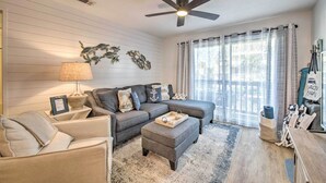 A living room with a sectional sofa, a small ottoman, a ceiling fan, and a large window with striped curtains. - PCB Condo Rental w/ Pool Access, Steps to Beach! (Panama City Beach)