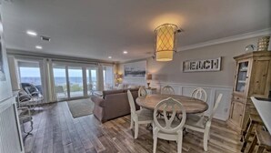 A spacious living area with a wooden floor, a round dining table with white chairs, a sofa, and a large window offering a view of the sea.