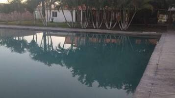 Une piscine rectangulaire dont l’eau reflète clairement les arbres et un bâtiment blanc situé à l’arrière-plan.