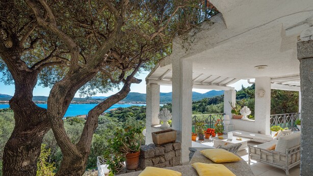 A covered outdoor seating area with stone seating and yellow cushions, overlooking a scenic landscape with trees and a view of the sea. - Apartment 'Bella Vista Marinella 1' with sea view, terrace, air conditioning and Wi-Fi (Olbia)