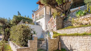 Un edificio de paredes de piedra con entrada porticada, un balcón con barandilla metálica y vegetación circundante.
