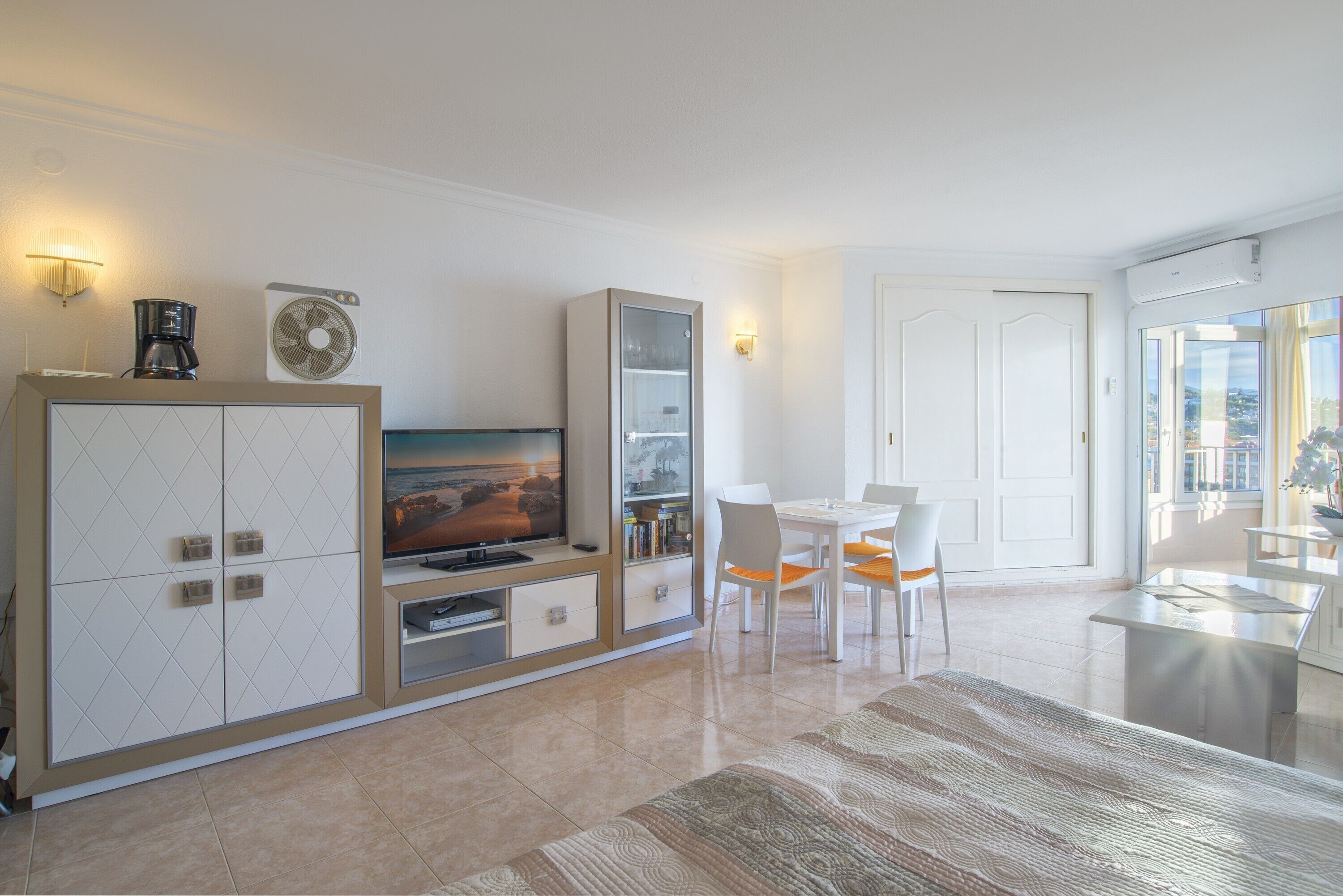 Een moderne woonkamer met een flatscreen televisie, een koffiezetapparaat, een ventilator en een eethoek met stoelen.