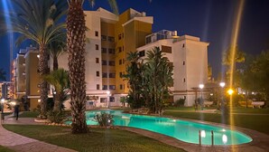 A nighttime view of a hotel with a swimming pool, palm trees, and a well-lit walkway.