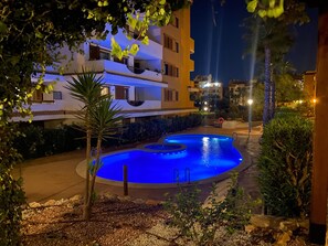 Zona de piscina nocturna con una piscina iluminada en azul, rodeada por un jardín ajardinado y un edificio con balcones.