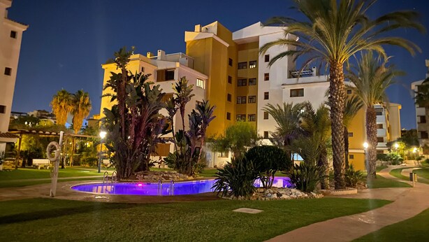 A nighttime view of a hotel with a swimming pool, palm trees, and a well-lit pathway.
