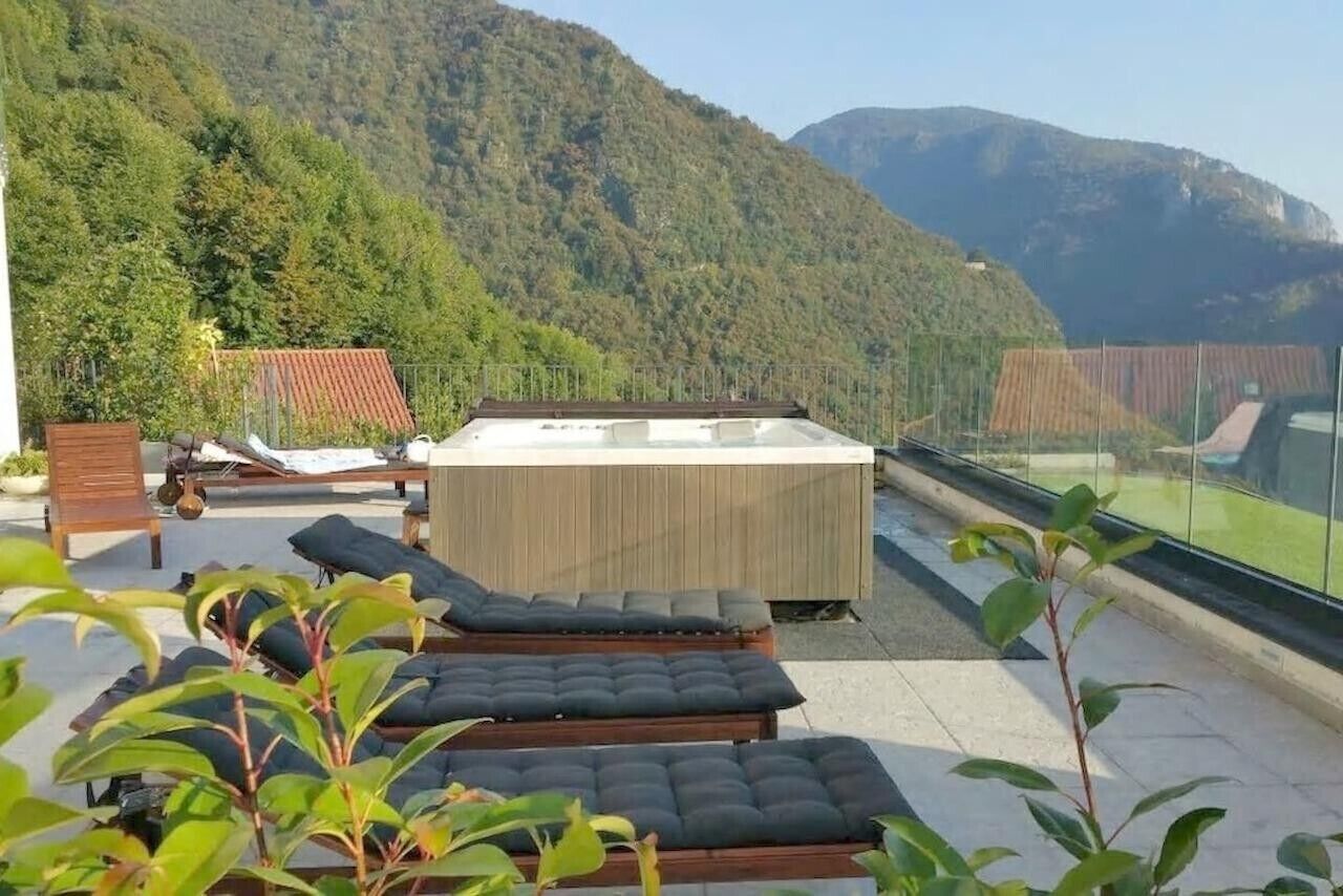 A rooftop terrace with lounge chairs, a hot tub, and a view of a forested mountain.
