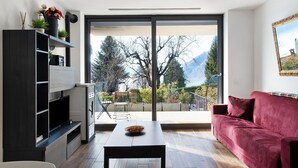 Un salon moderne doté d’un téléviseur à écran plat, d’un canapé rouge, d’une table à manger et d’un balcon offrant une vue sur les arbres et les montagnes.