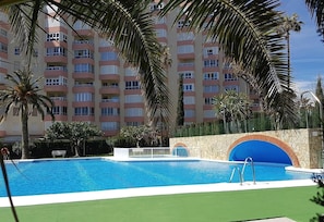 A swimming pool with a stone wall and a blue arch, surrounded by palm trees and a multi-story building.