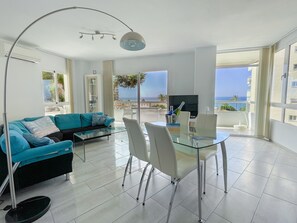 A modern living room with a glass table, white chairs, a blue sofa, and a large TV. - Apartment Front Americano Near the Beach with Sea View, Pool (Torrox Costa)