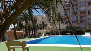 En pool med en bänk längs kanten, omgiven av palmer och en byggnad med flera våningar.