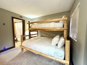Un lit superposé avec une structure en bois et une literie blanche, dans une pièce dont le sol est recouvert d’une moquette et qui possède une fenêtre avec des stores.