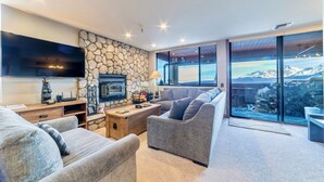 Un salon moderne avec un mur en pierre, un téléviseur à écran plat, une cheminée et une grande fenêtre donnant sur une vue imprenable sur les montagnes.