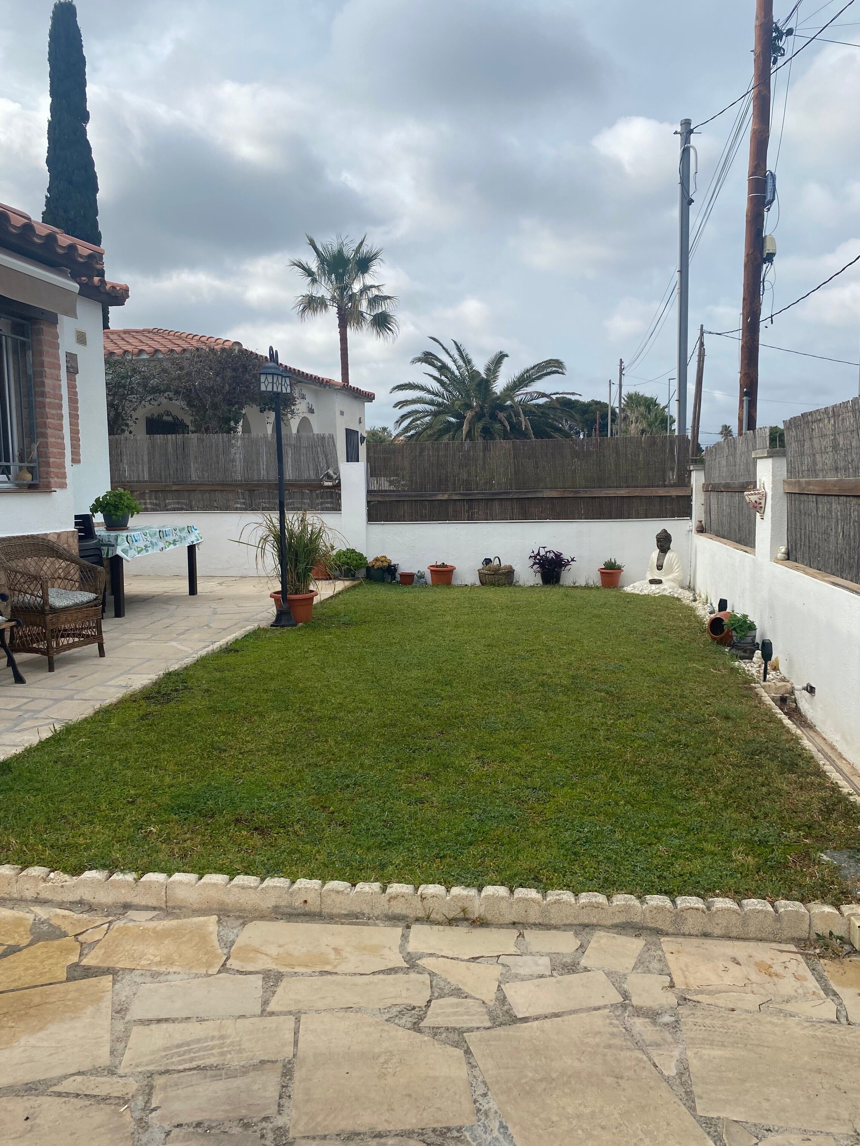 Un patio con césped, plantas en macetas, una silla de mimbre y un sendero de piedra.