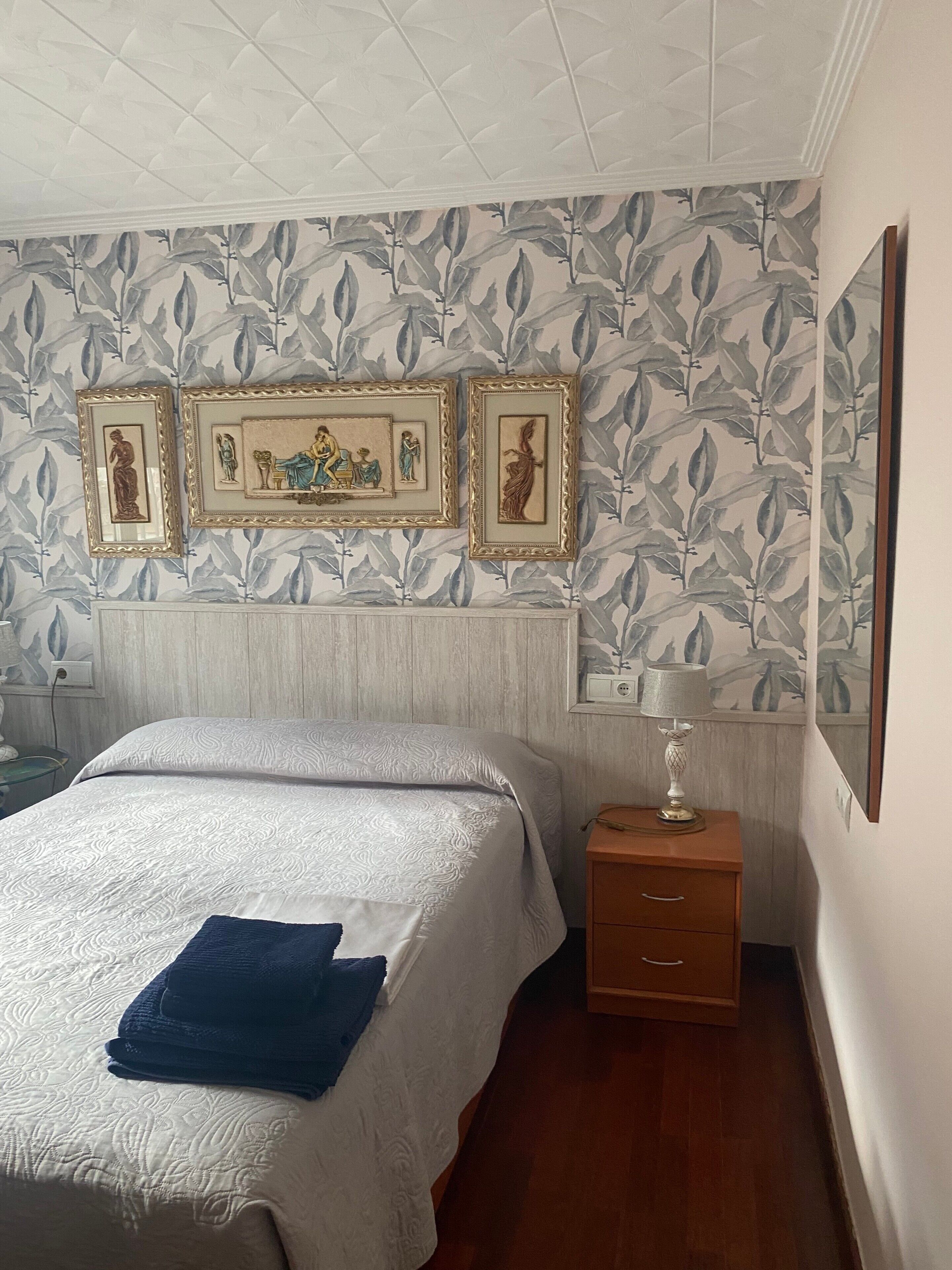A neatly made bed with a blue towel, a wooden nightstand with a lamp, and three framed artworks on the wall.