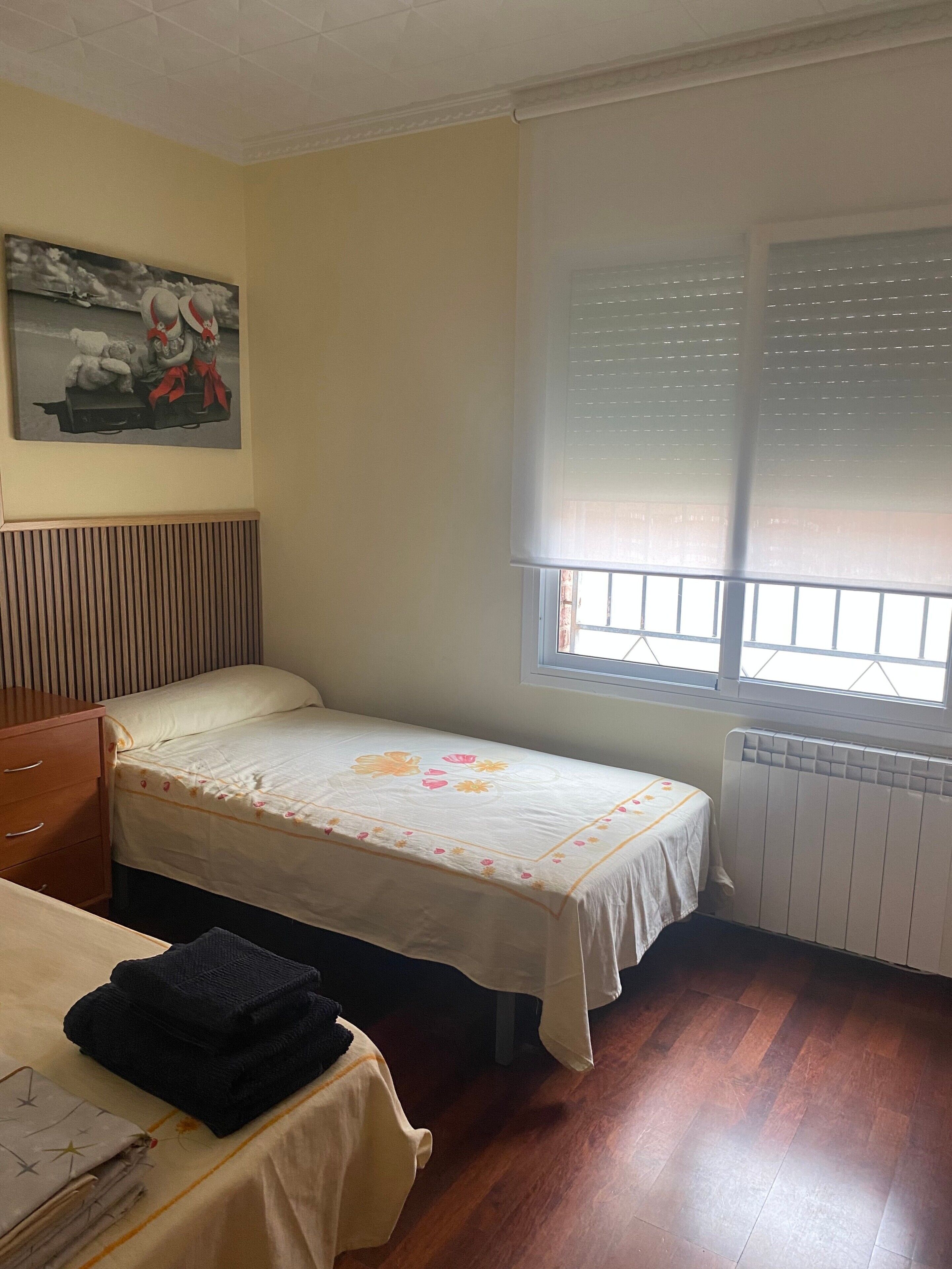 A small bedroom with a single bed, a wooden dresser, a radiator, a window with blinds, and a painting on the wall.
