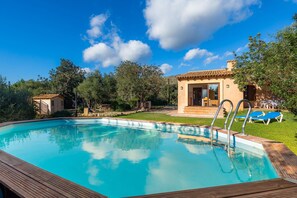 Una piscina con muelle de madera, una casa pequeña con techo de tejas y un área verde con sillas de descanso.