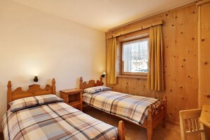 Ein Zimmer mit zwei hölzernen Betten, einem kleinen Holztisch, einem Stuhl, einem Fenster mit Vorhängen und einer Wandleuchte.